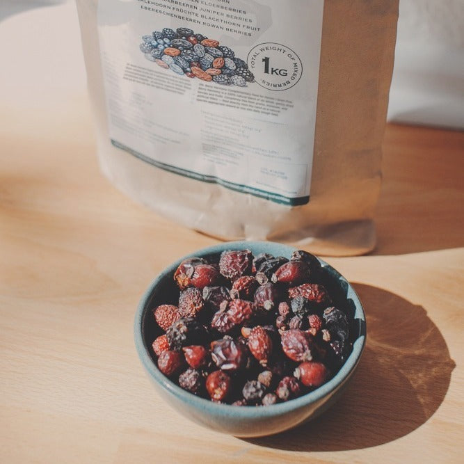 Ceramic bowl filled with whole dried rosehips, hawthorn, elderberries, juniper, sloe, and rowan berries in front of the 1 kg kraft paper bag